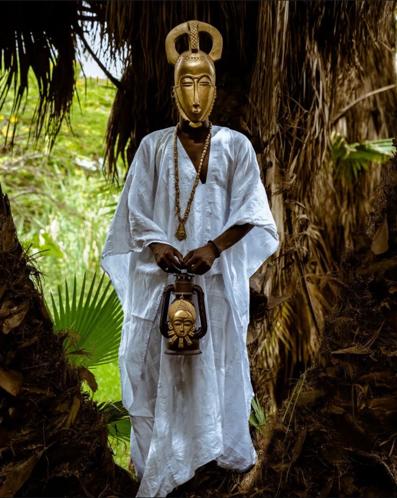A figure in a white robe holding a carved mask, standing among palm fronds